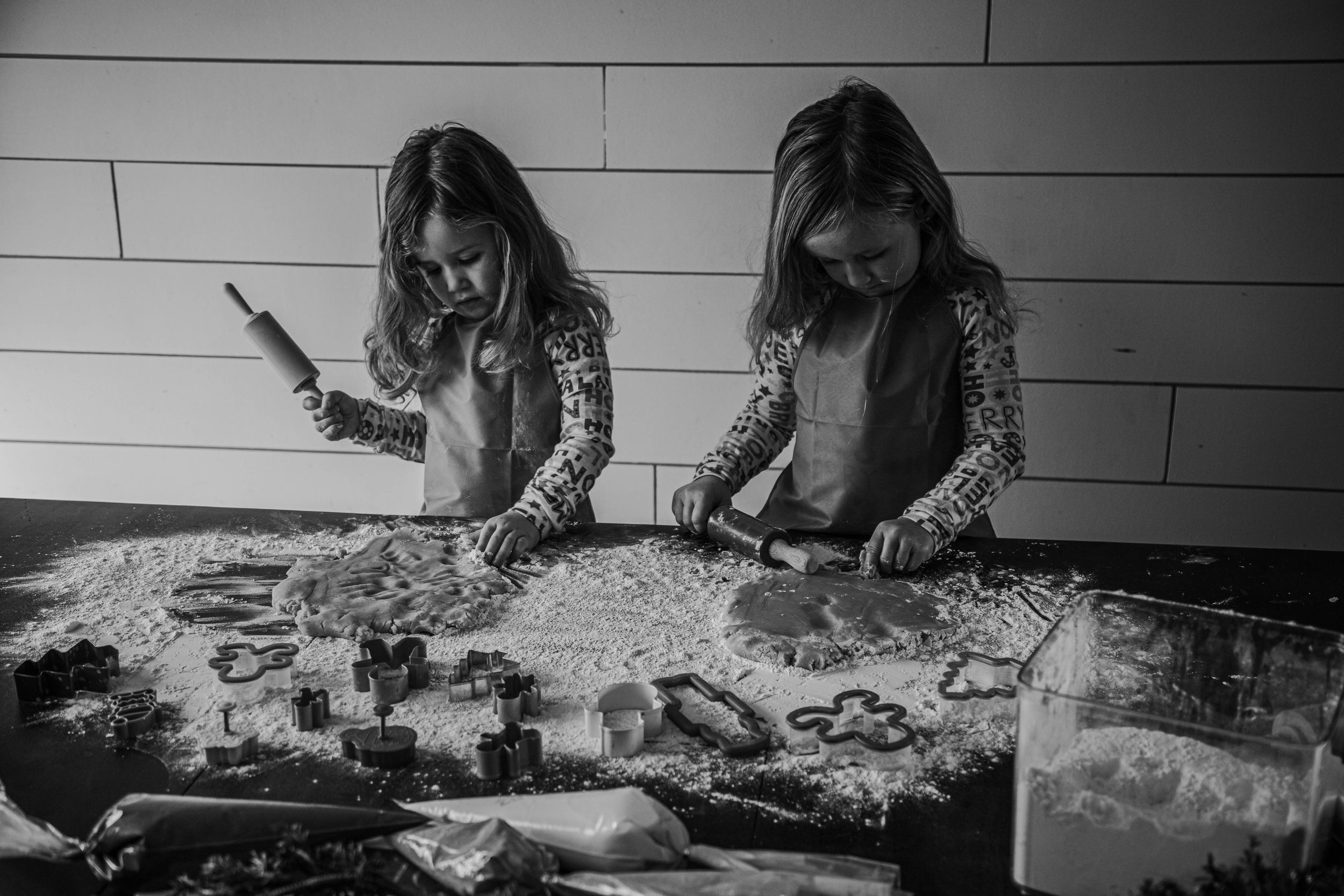 Two young girls in aprons rolling dough and using cookie cutters on a flour-covered kitchen counter, black and white.