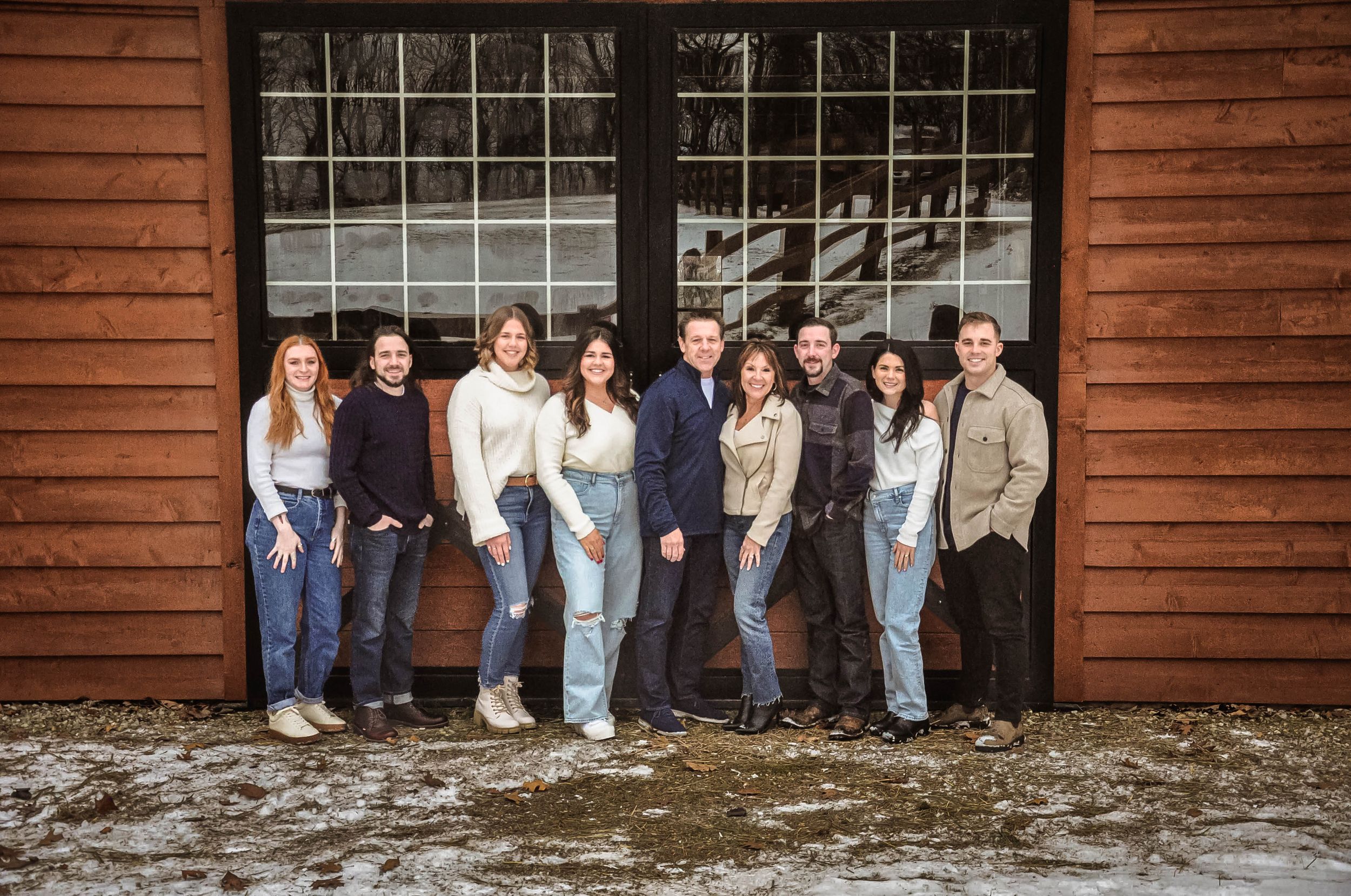 Group of nine adults posing and smiling in front of a rustic wooden barn door in a snowy winter setting.