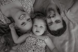 Black and white overhead portrait of parents and baby lying together smiling.