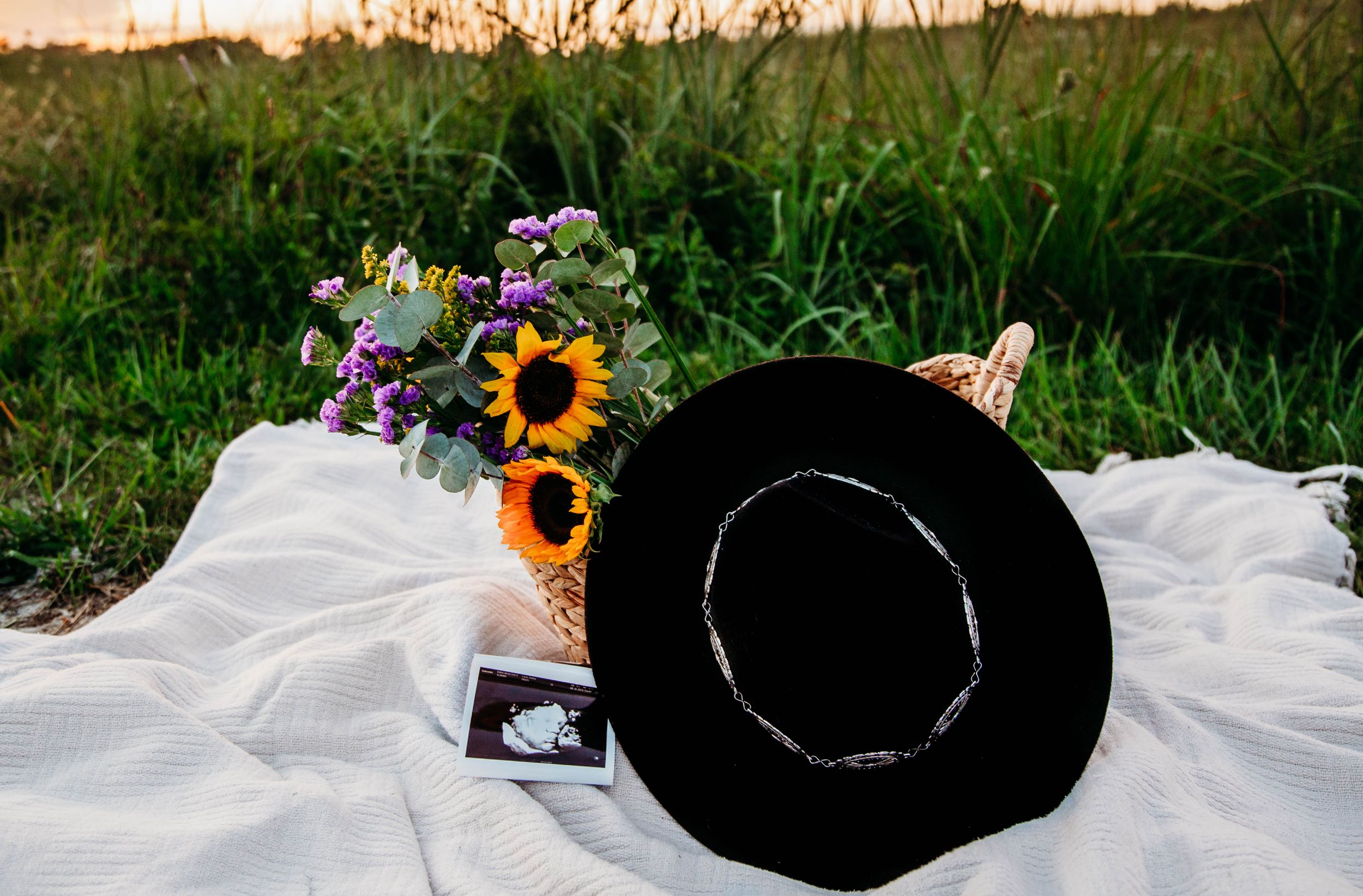 Black wide-brim hat and sunflower bouquet with ultrasound photos on white blanket in a field at sunset.