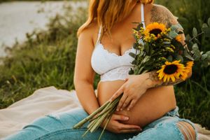 Pregnant woman in white bralette holding sunflowers while sitting in grass.
