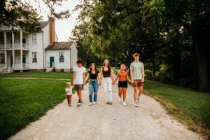 Family of six walking hand-in-hand down a gravel path near a historic white house.