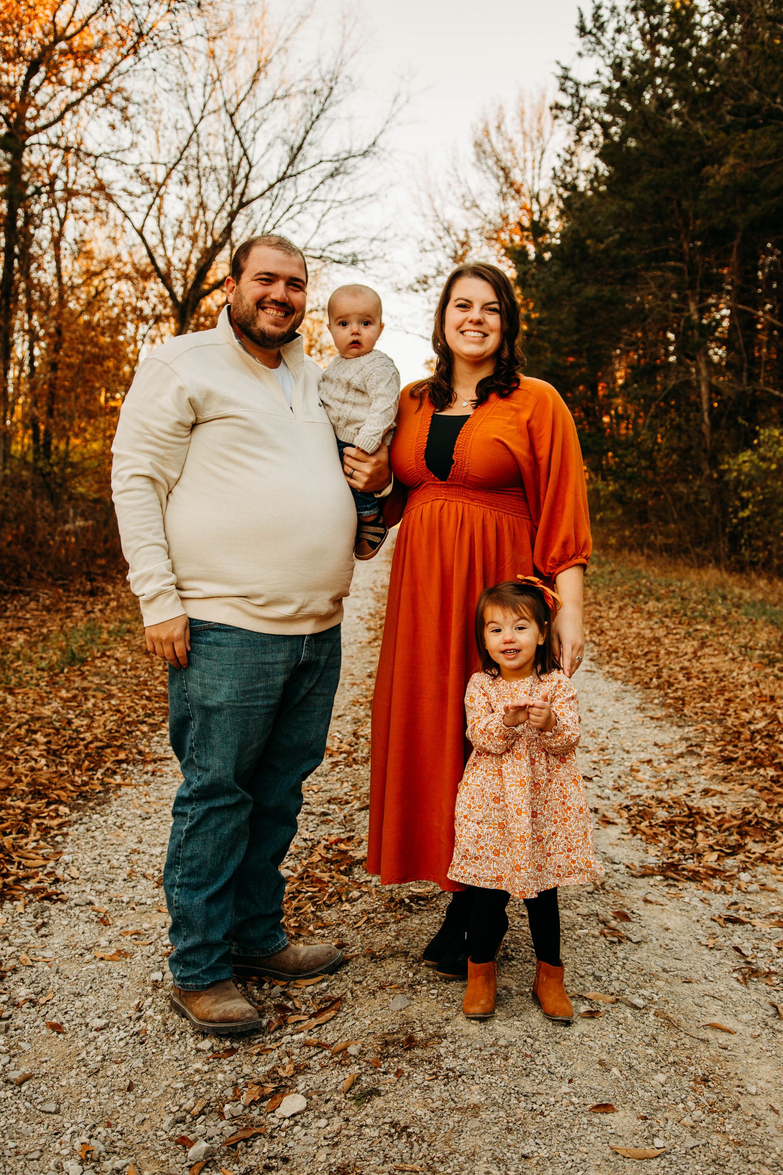 Family of four posing on an autumn leaf-covered path, mother in orange dress holding baby, young girl standing nearby.