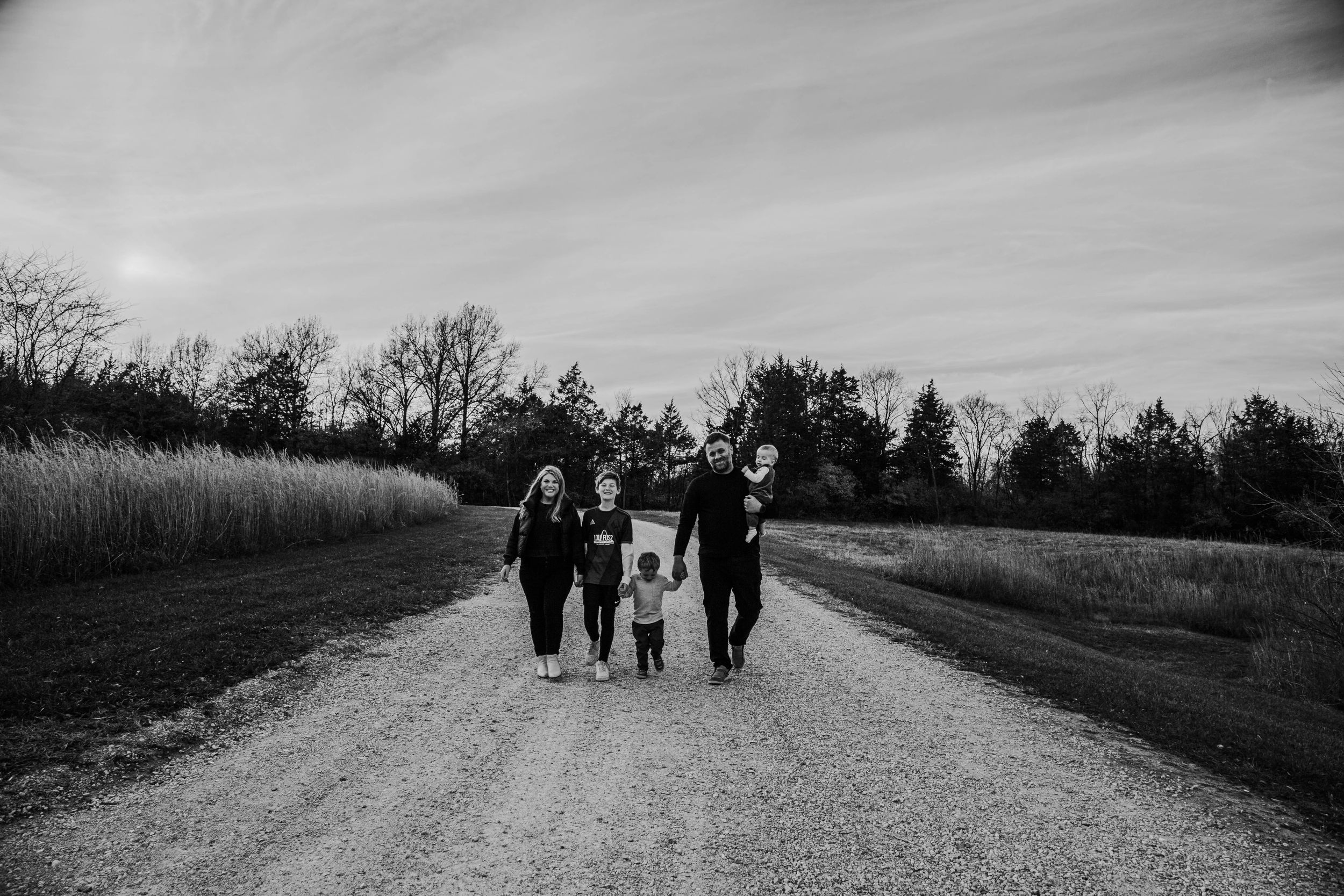 Black and white photo of a family of four walking hand-in-hand down a rural gravel road surrounded by trees.