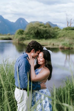 A romantic couple shares tender moments by a serene lake with mountain views and tall grass in the background.