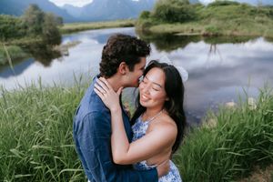 Couple embraces tenderly by a serene mountain lake with tall grass and reflective water in the background.