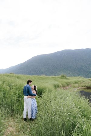 Couple embraces in a scenic mountain meadow with tall green grass and rolling hills in the background.