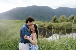 A couple embraces in a grassy meadow with mountains and a lake in the scenic natural backdrop.