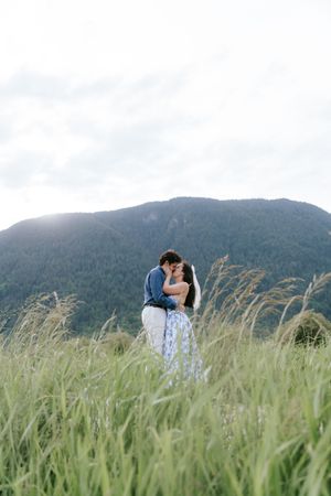A couple embraces in a field with tall grass and mountains in the background on an overcast day.
