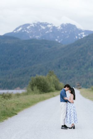 Two people share a romantic moment on a mountain road with snow-capped peaks and forested hills in the background.