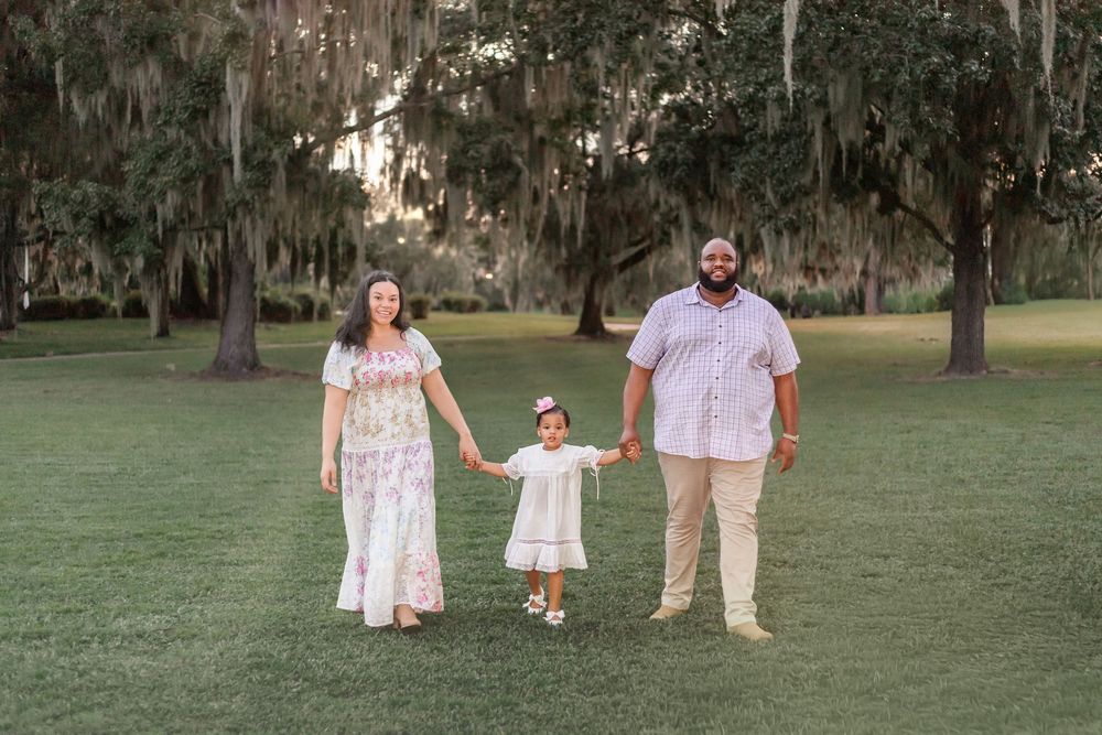 A family stands together holding hands in a grassy park with Spanish moss draped oak trees in the background.