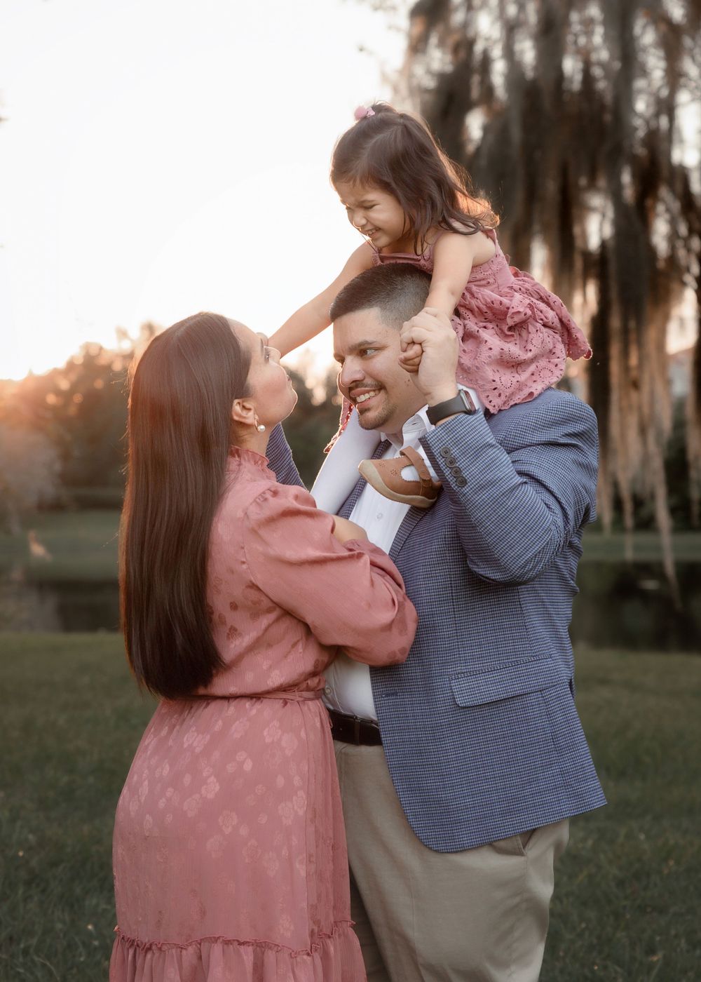 A couple in pink and blue attire shares joyful moments together during a sunset photoshoot in a grassy field.