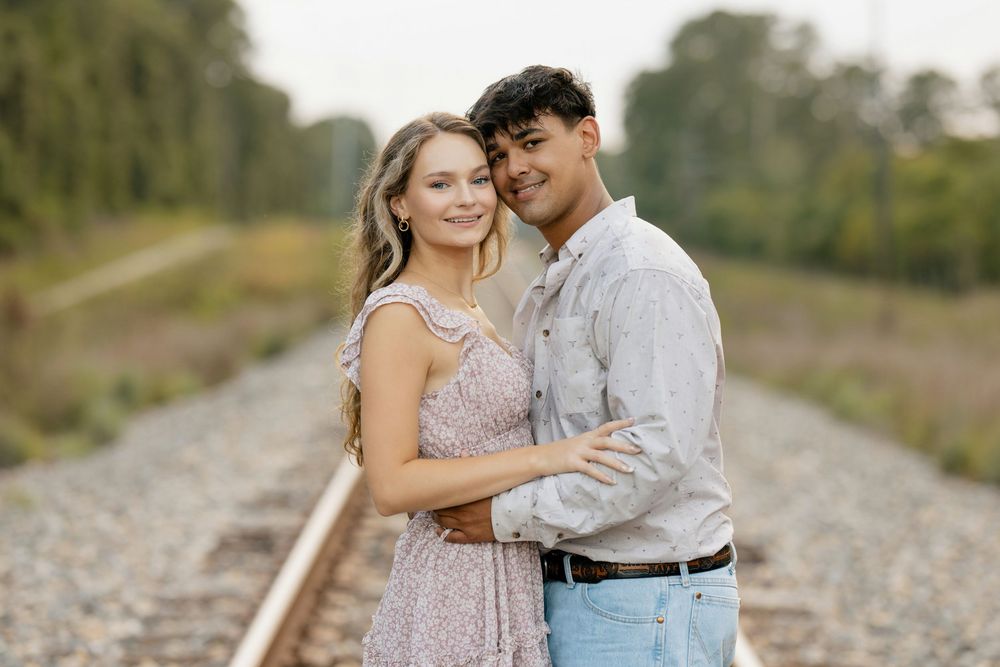A romantic couple embraces on a railroad track at sunset wearing light colored clothing.
