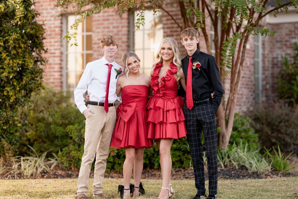 Formal group photo of friends in red dresses and suits posing together outdoors against a brick wall backdrop.