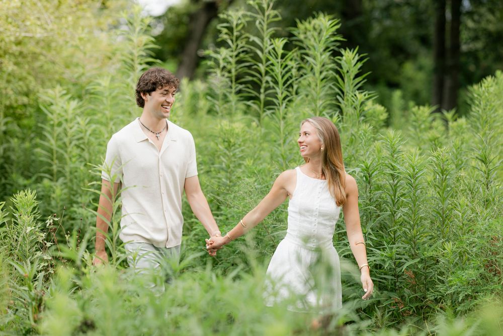 A couple holds hands while walking together through tall green plants on a sunny summer day.