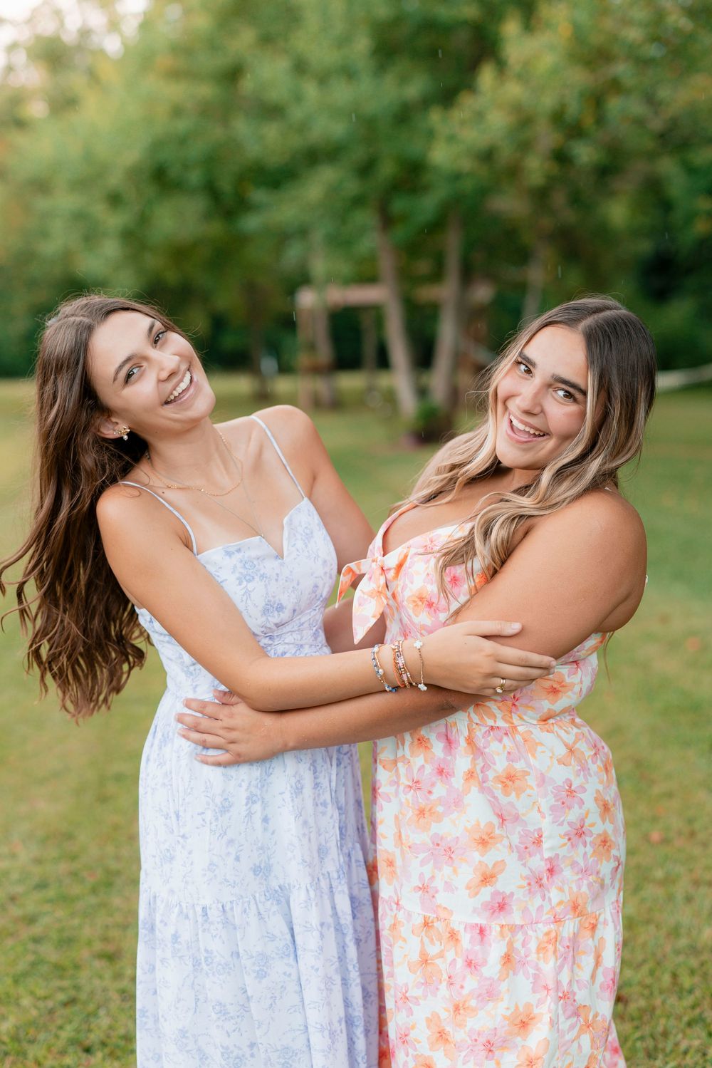 Two friends wearing summer dresses share candid laughs and hugs in a sunlit outdoor garden setting.