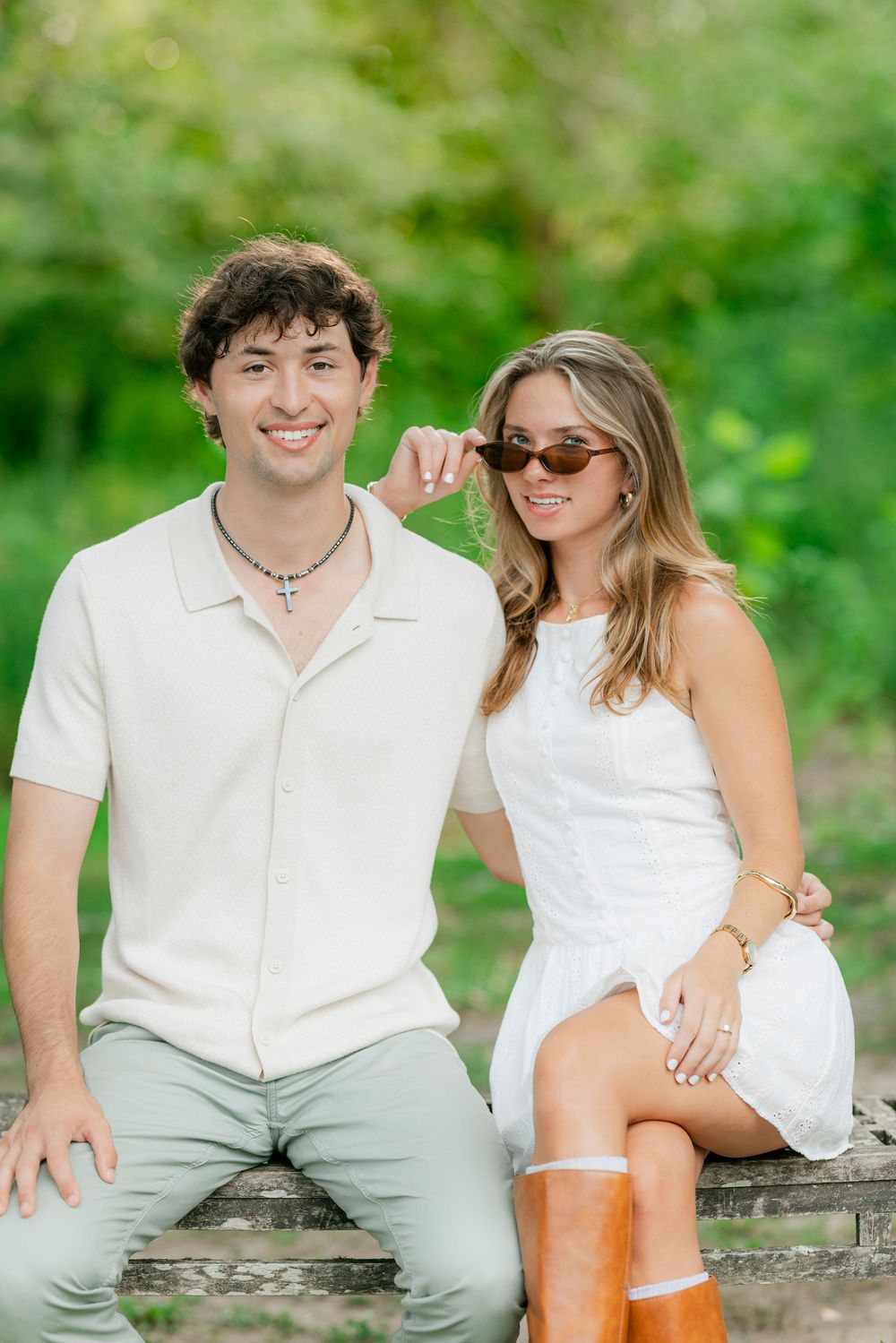 A couple in coordinating white outfits pose together along a rustic wooden platform in an outdoor garden setting.