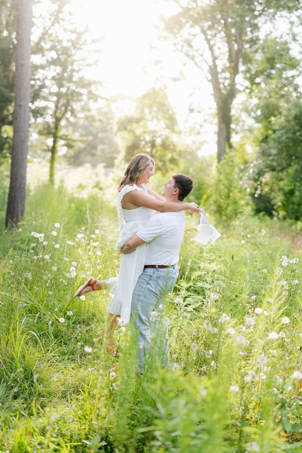 A couple shares a romantic moment dancing together in a sunlit meadow filled with wildflowers and tall grass.