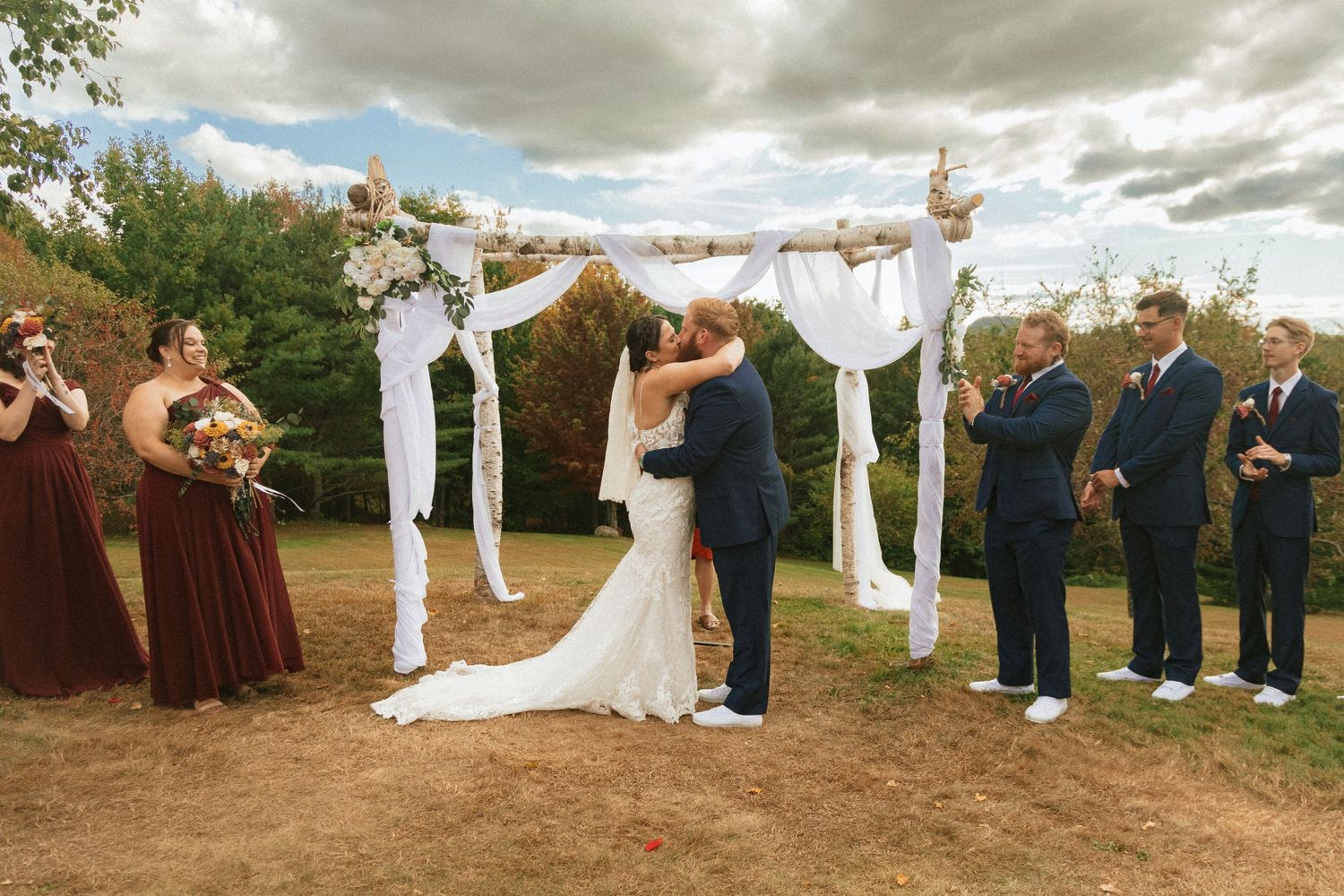 A wedding ceremony takes place outdoors under a white draped arch in Maine with wedding party in burgundy and navy attire.