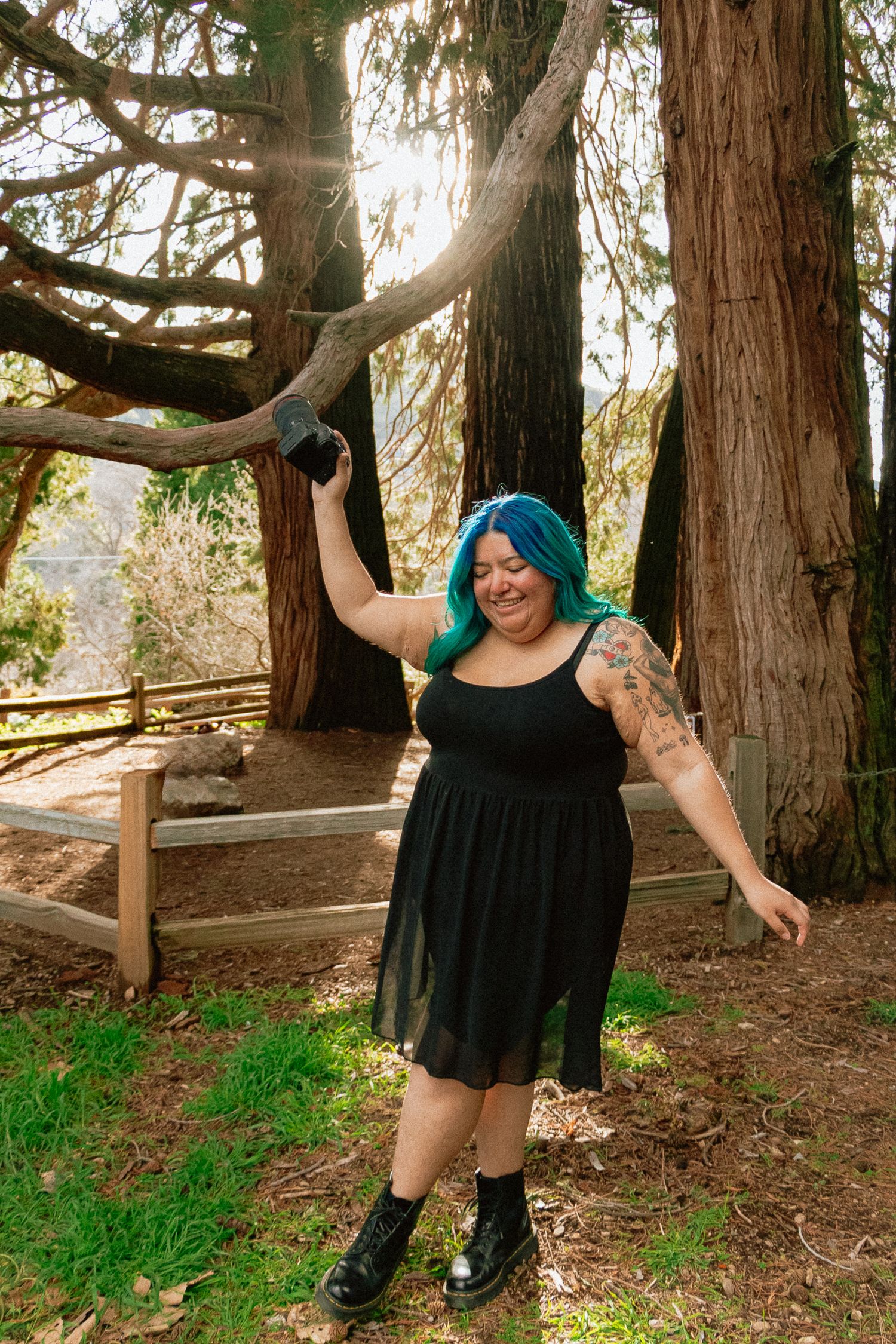 Mexican woman Photographer with bright turquoise hair and tattoos holds professional camera while smiling in forest setting.