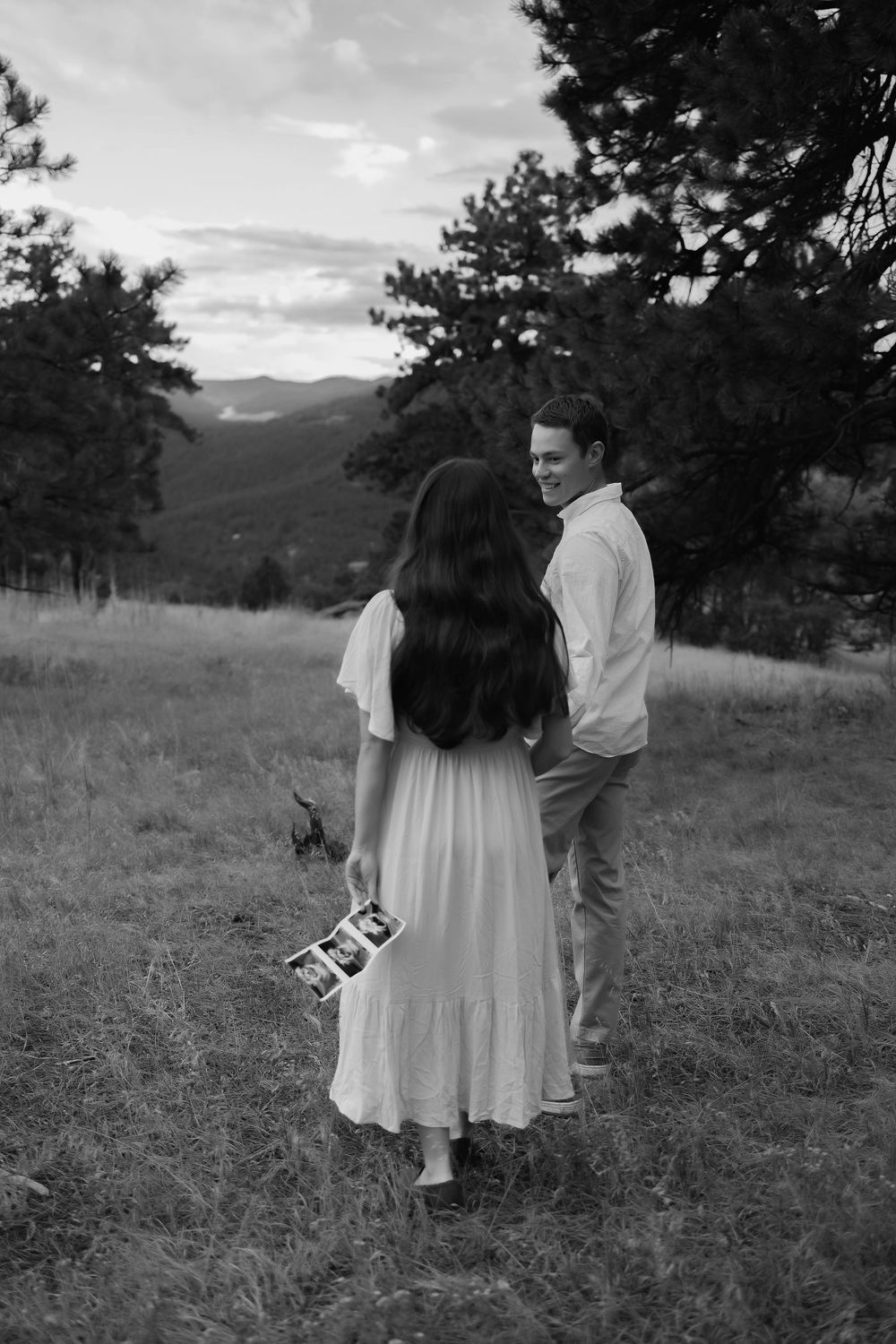 Black and white photo of a couple standing in a mountain meadow, woman in white dress holding ultrasound photos.