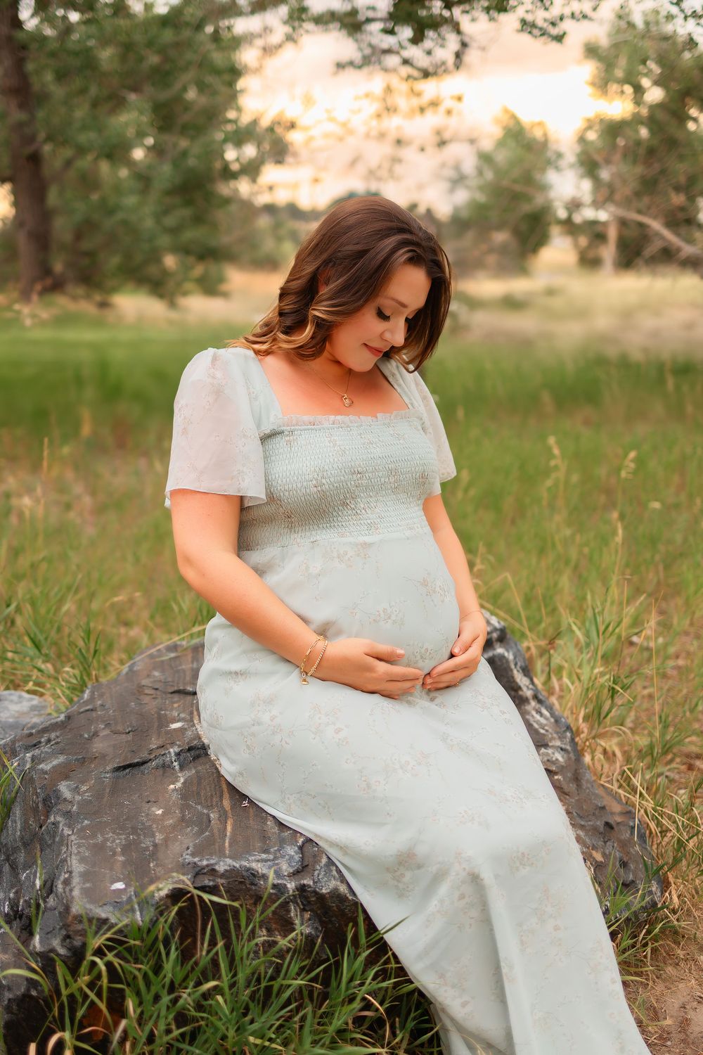 Pregnant woman in mint green dress sitting on rock in golden hour field, cradling baby bump outdoors.