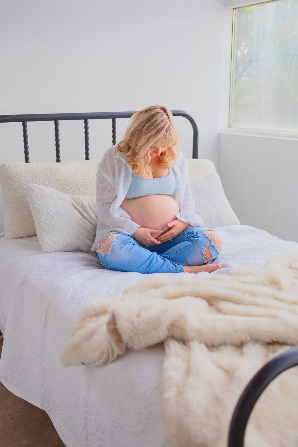 Pregnant woman in ripped jeans and blue crop top sitting cross-legged on bed, cradling baby bump in bright bedroom.