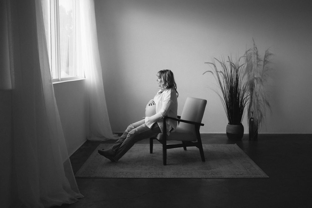 Black and white photo of a pregnant woman sitting in a chair by a window, gazing thoughtfully in a minimalist room.