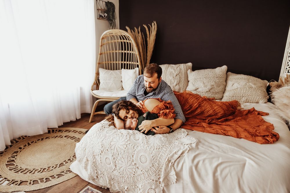 Couple relaxing together on bed with white and rust bedding near a rattan chair by window. Snuggling their newborn baby
