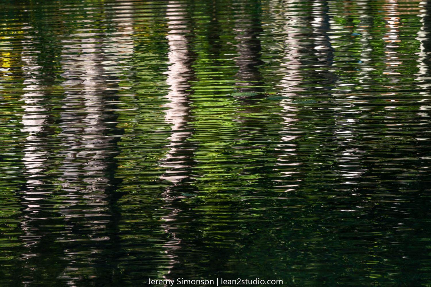 Abstract reflection of trees creating vertical green and white striped patterns on water.