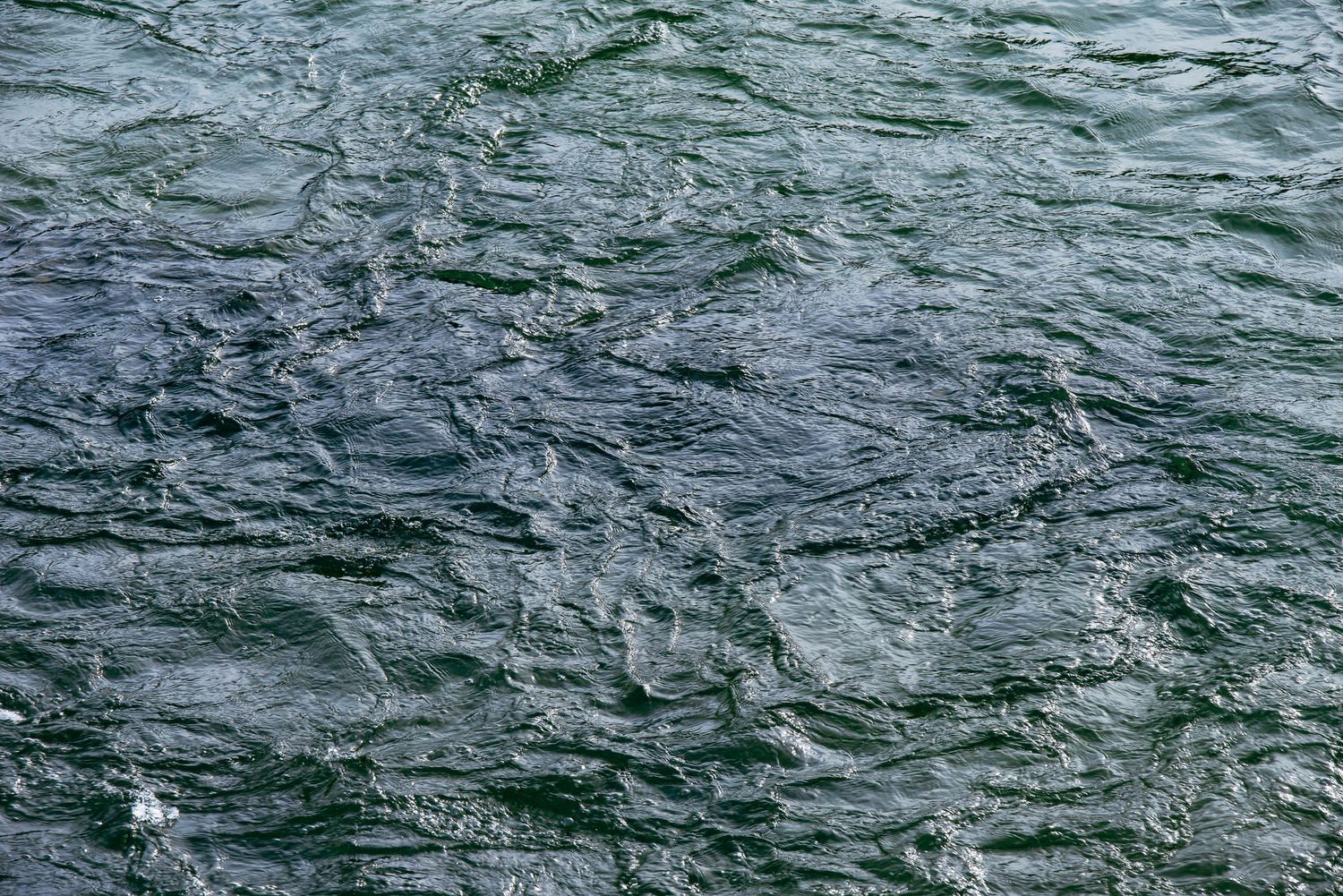 Rippling dark blue-green water surface with textured waves and light reflections creating an abstract pattern.