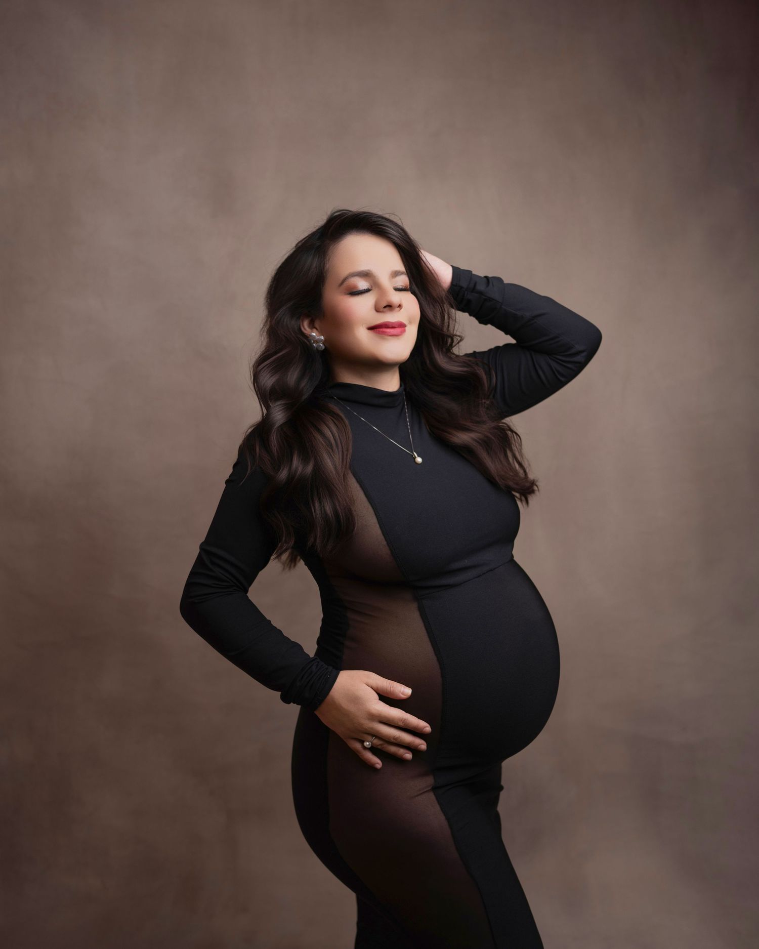 A pregnancy portrait in a black fitted dress against a brown background showing a serene pose with one hand raised.