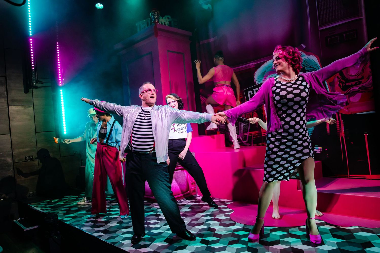 Dynamic dance scene on stage with performers in retro costumes under vibrant pink and blue lighting.