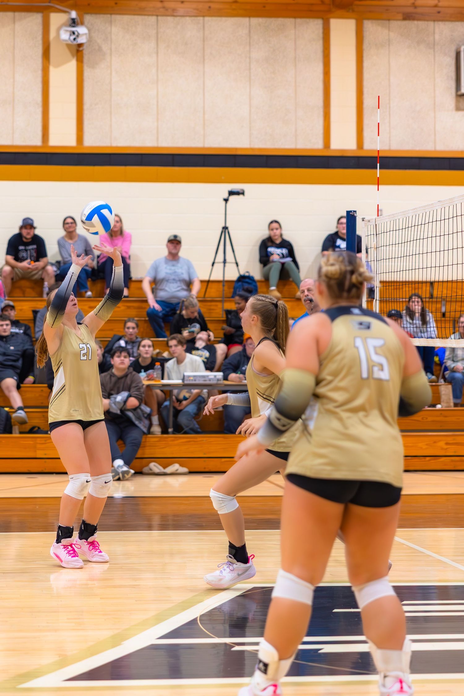 Volleyball players in gold uniforms diving for a ball during an indoor match on a wooden court.