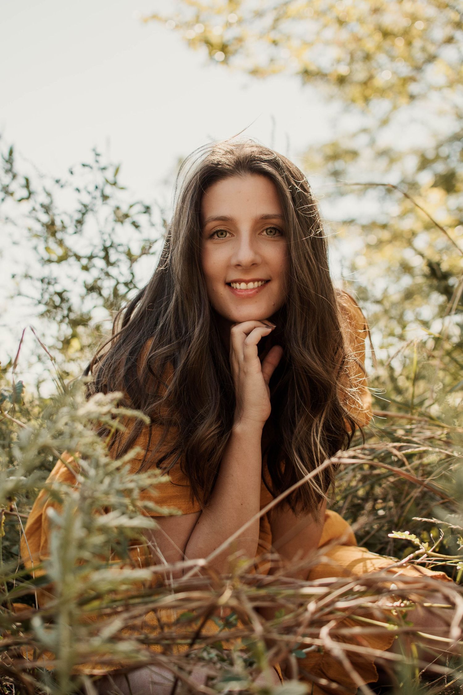 Woman in yellow dress and sun hat poses elegantly in tall dried grass at sunset.
