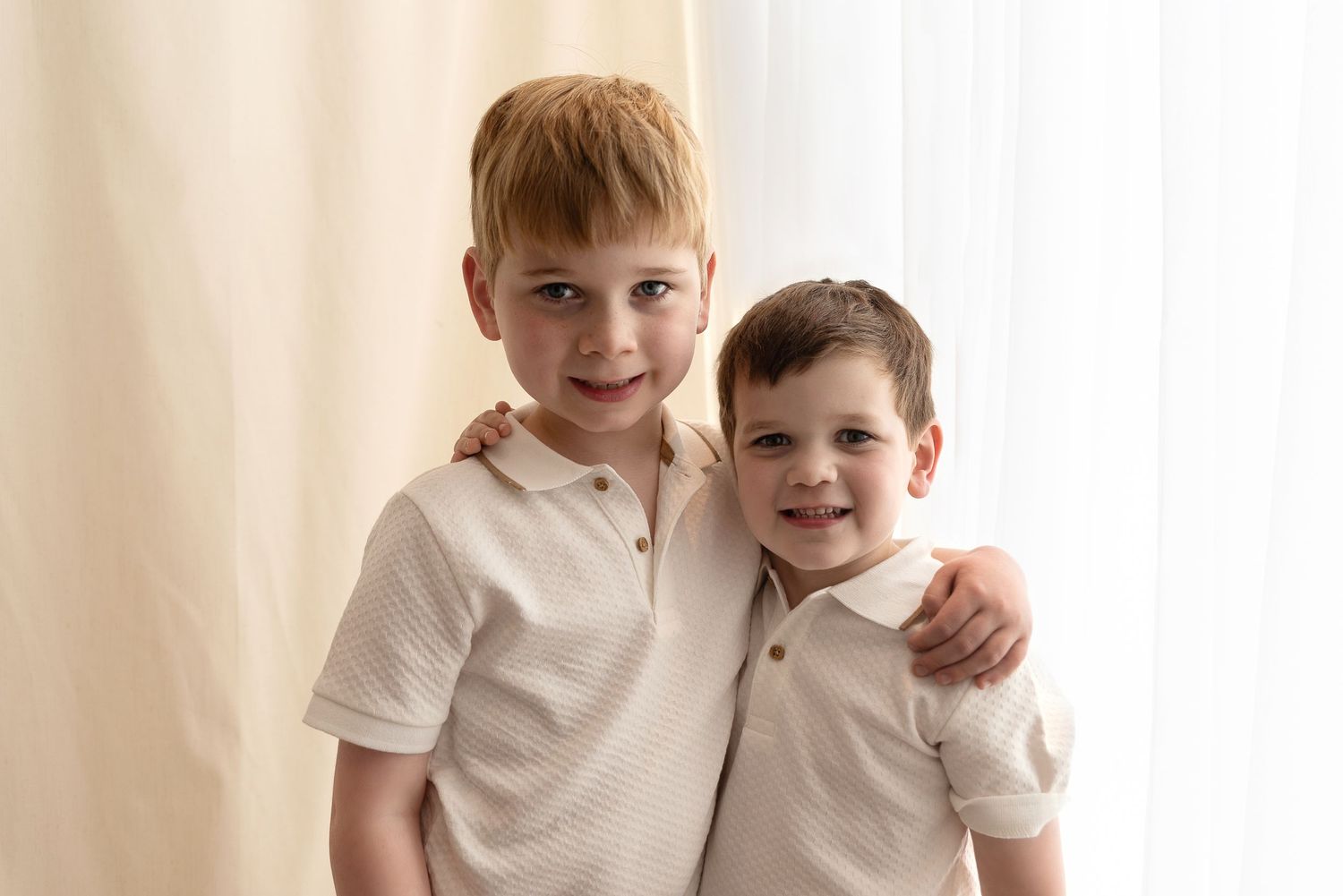 Two young children wearing white polo shirts stand together smiling against a light colored wall.