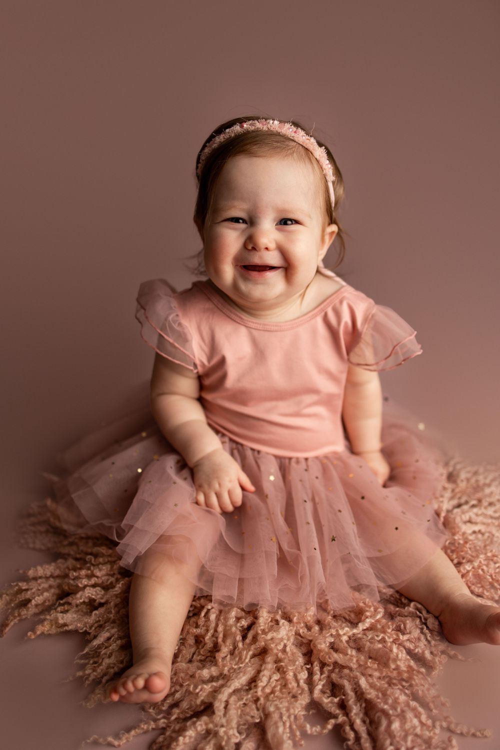 A baby wearing a pink tulle dress sits on a plush carpet in a studio setting, showing a bright happy expression.