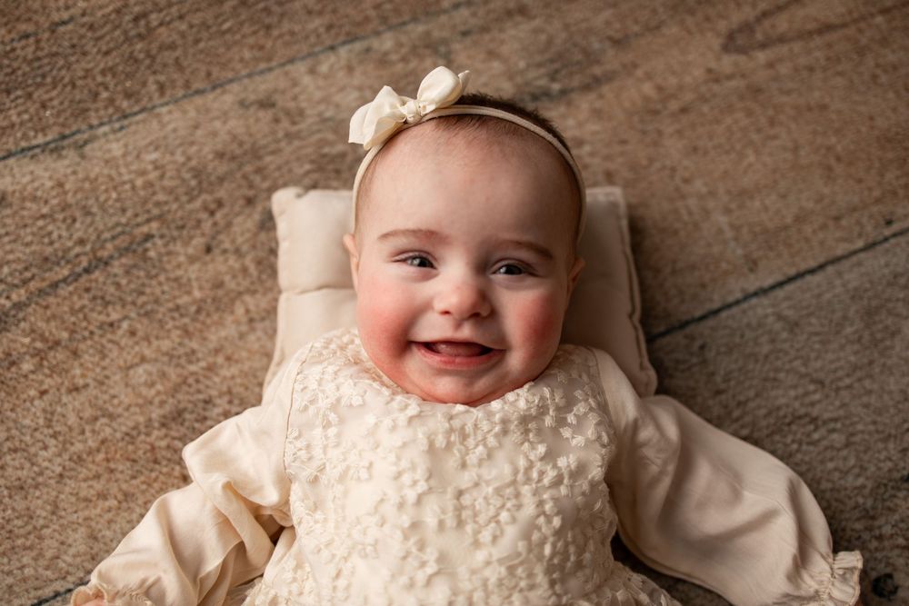 A baby in cream-colored clothing and bow headband smiles while lying on a wooden floor.