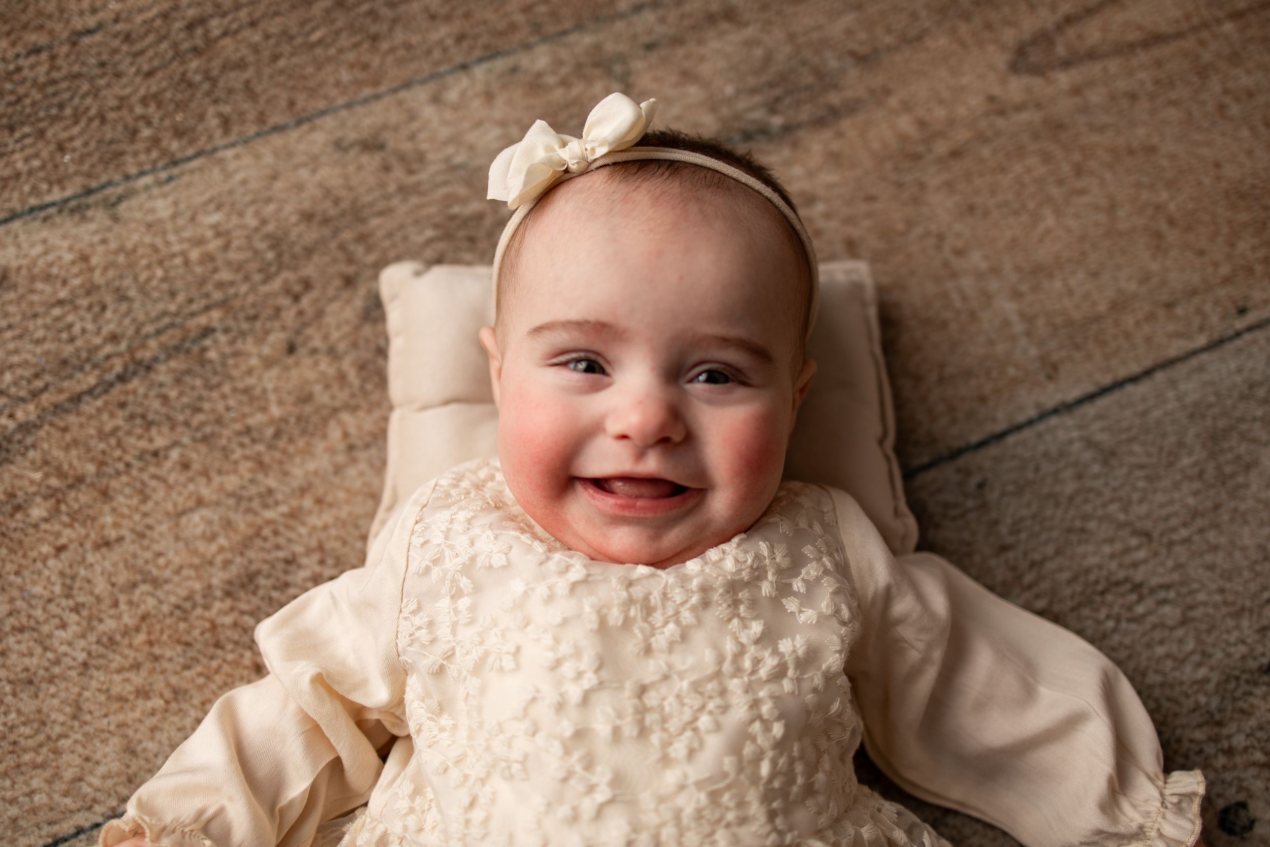 A baby in cream-colored clothing and bow headband smiles while lying on a wooden floor.