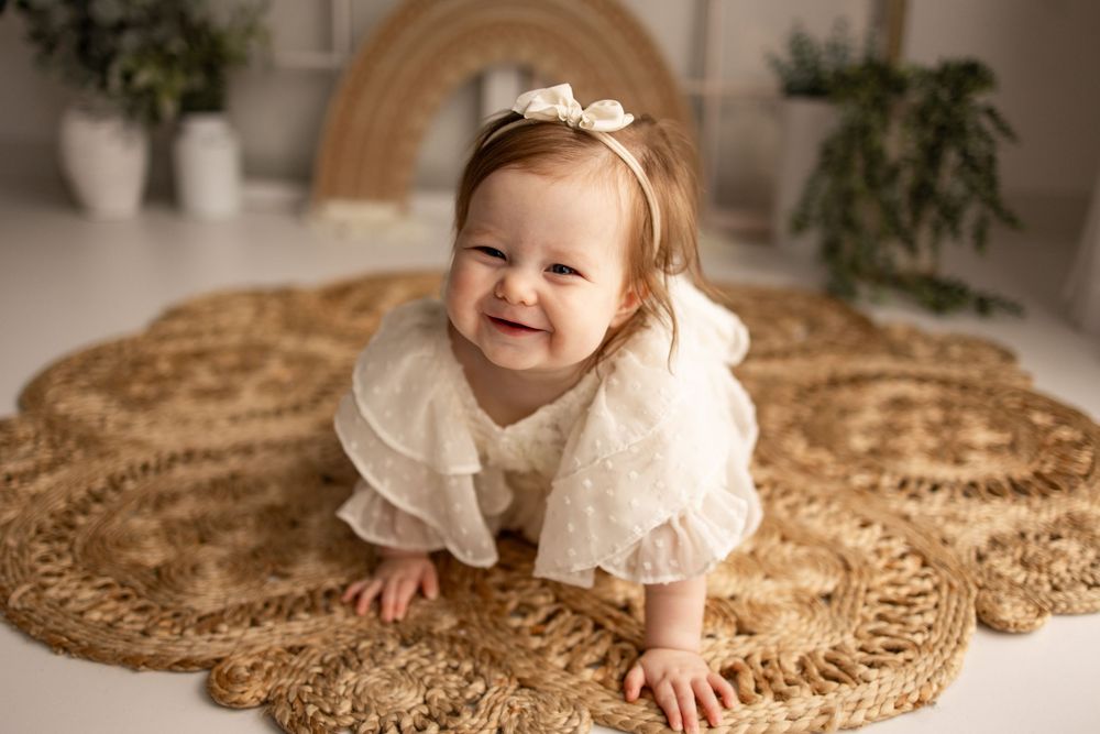 A baby in a white dress crawls on a round jute rug in a bright interior setting.