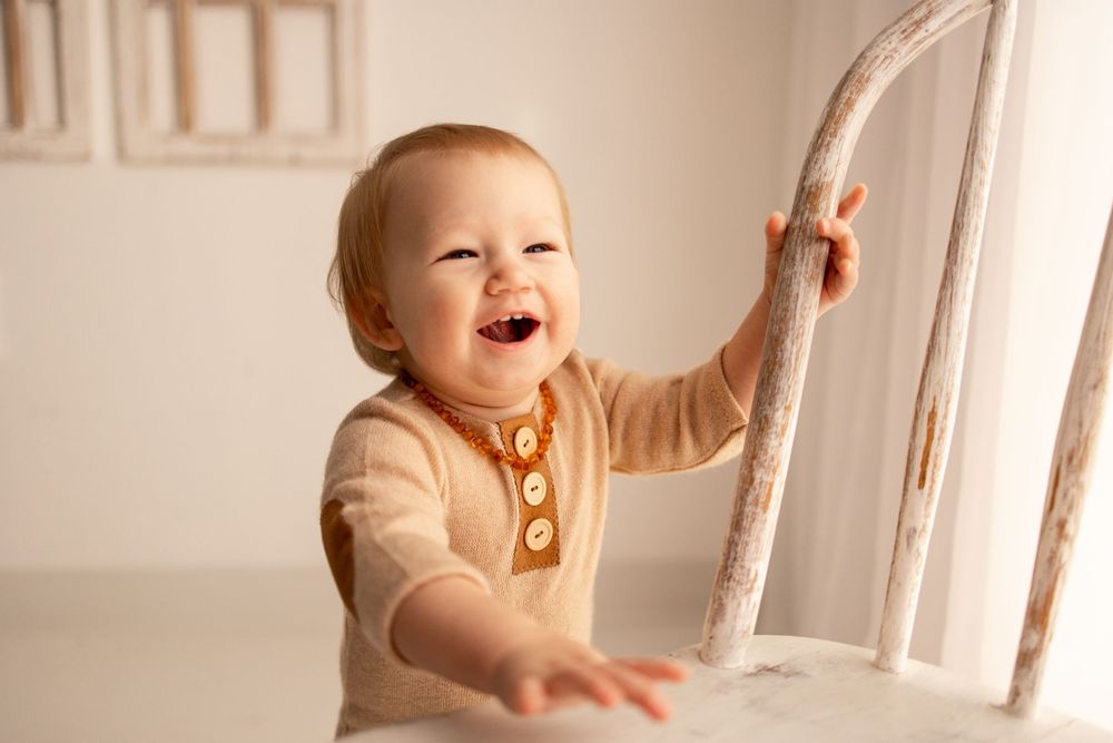 A happy baby wearing a white outfit laughs while standing and holding onto white wooden spindles of a staircase.