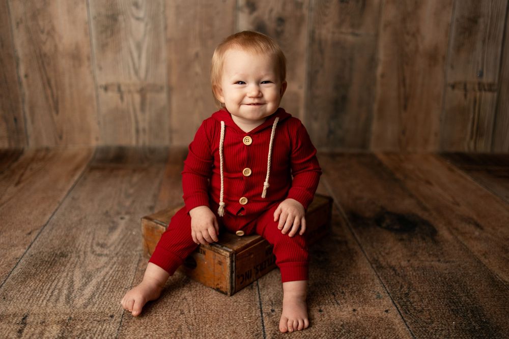 A small child wearing a red knit romper sits on a wooden floor smiling while resting on a vintage wooden box.