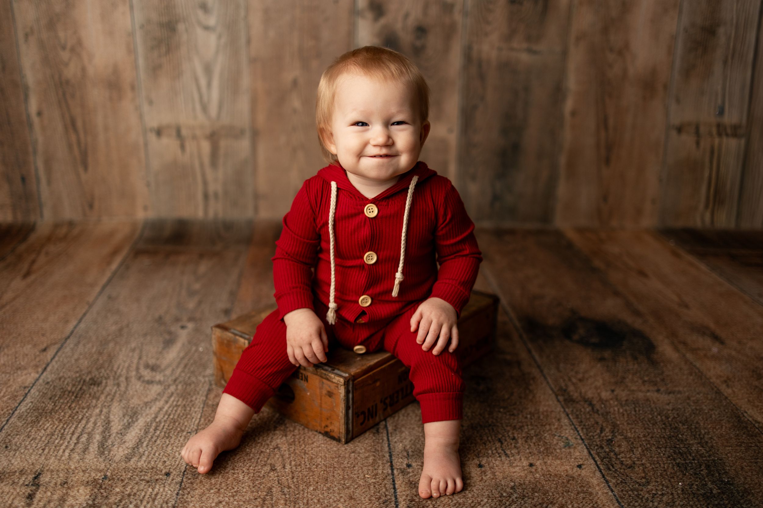 A small child wearing a red knit romper sits on a wooden floor smiling while resting on a vintage wooden box.