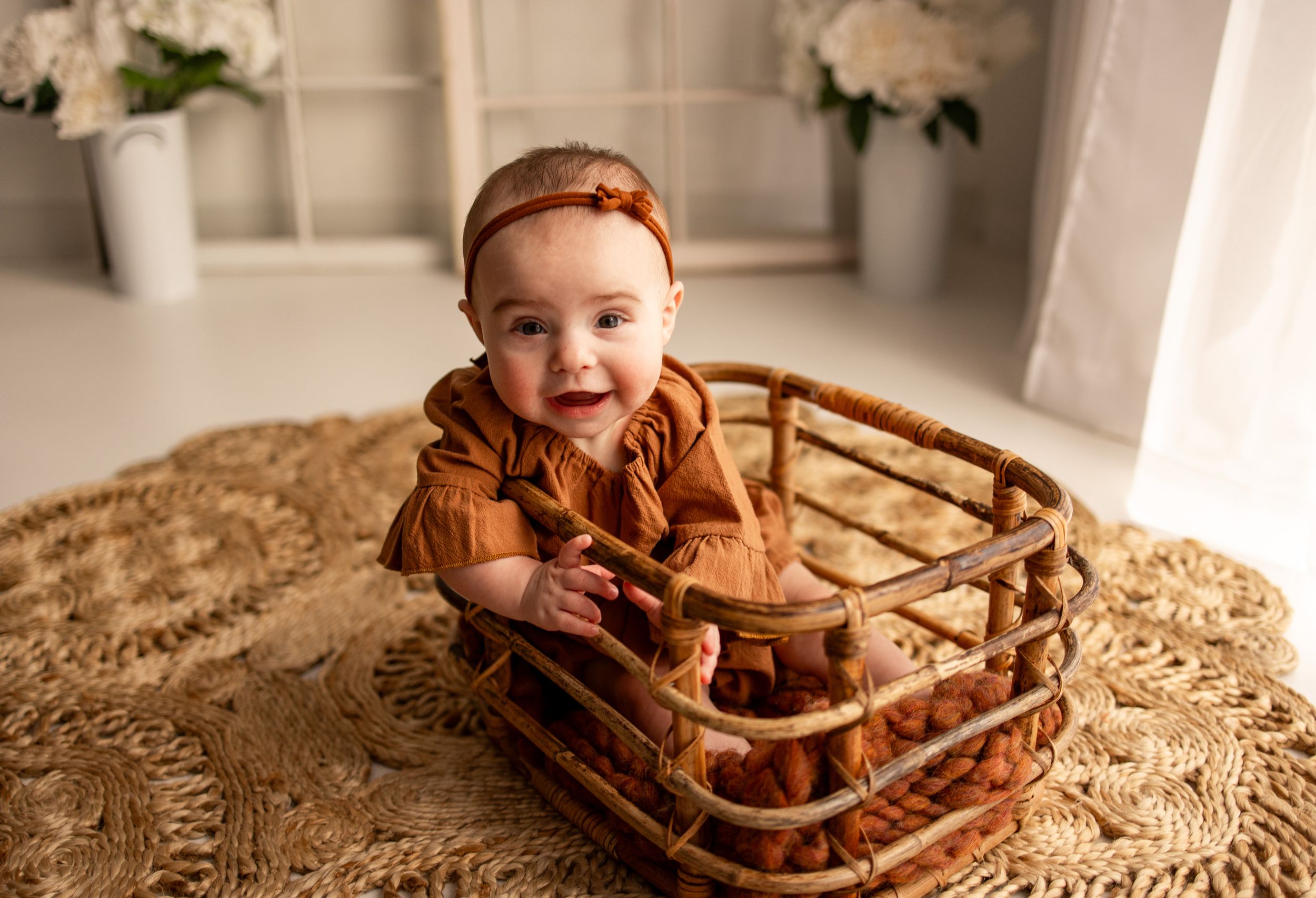 A baby sits in a wooden bowl on a textured beige blanket, smiling brightly at the camera.