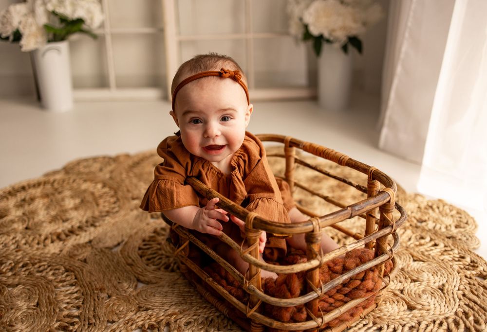 A baby sits in a wooden bowl on a textured beige blanket, smiling brightly at the camera.