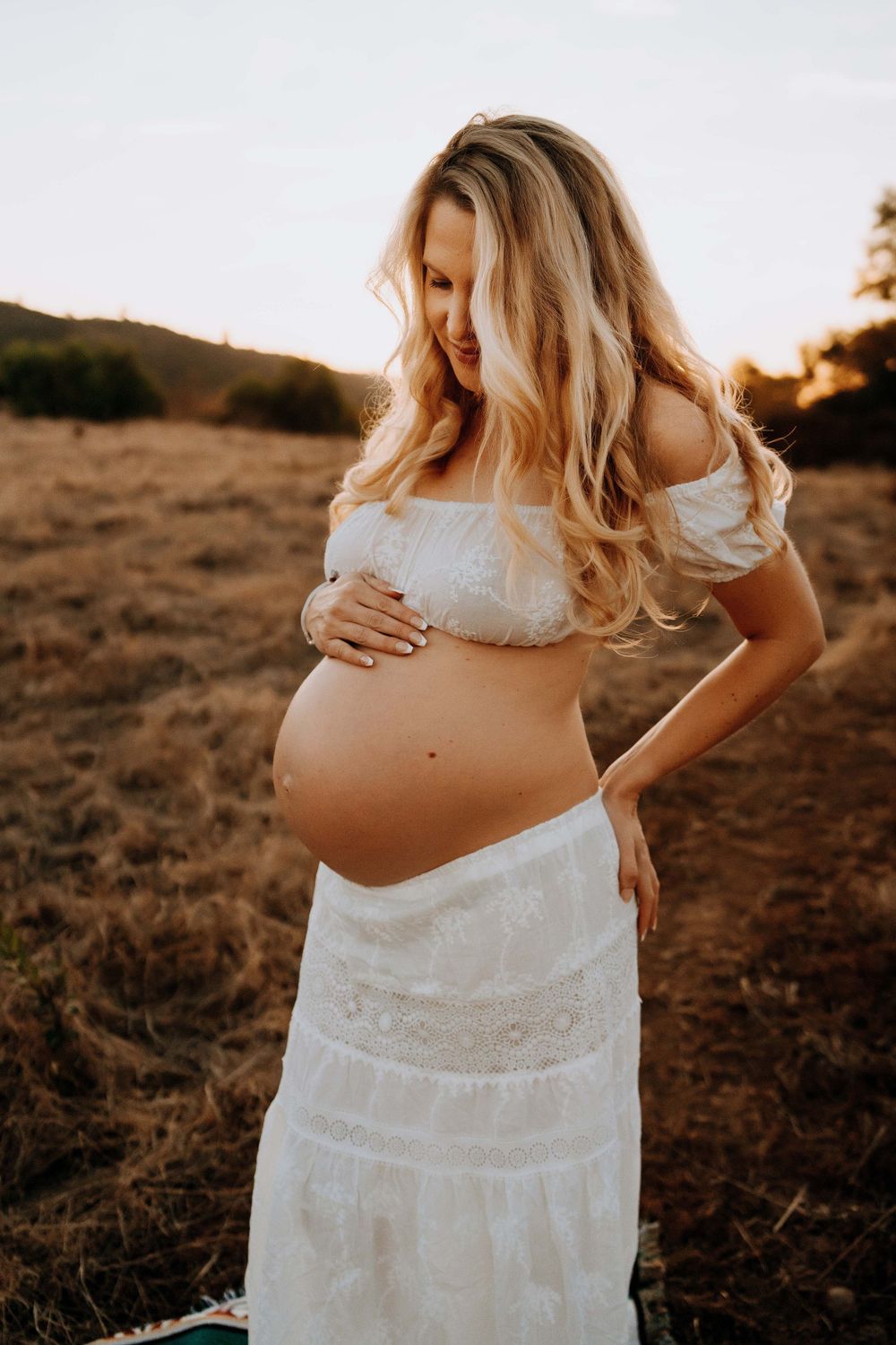 Pregnant woman in white off-shoulder crop top and skirt cradling baby bump in golden field at sunset.
