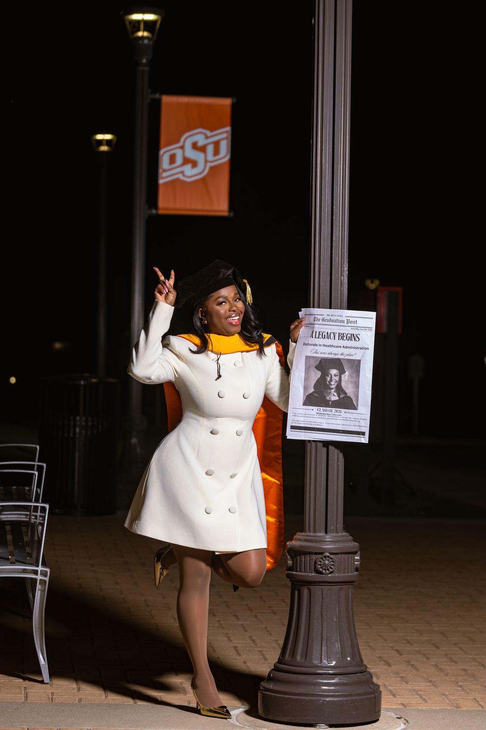 Woman in white coat poses by lamp post at OSU campus at night, making peace sign gesture.