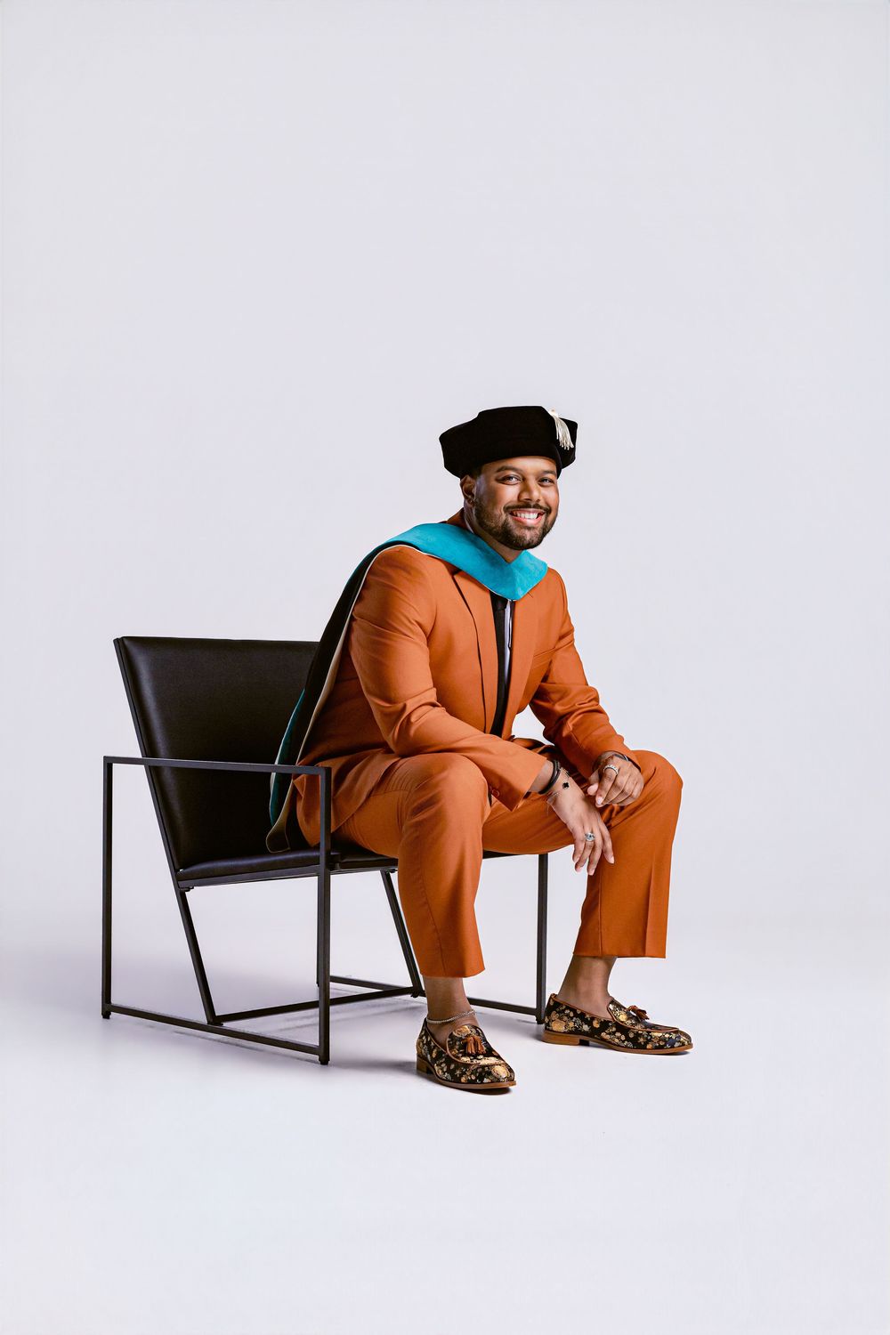 Man in orange suit and black doctoral cap sits smiling in modern chair, wearing academic regalia with teal hood.
