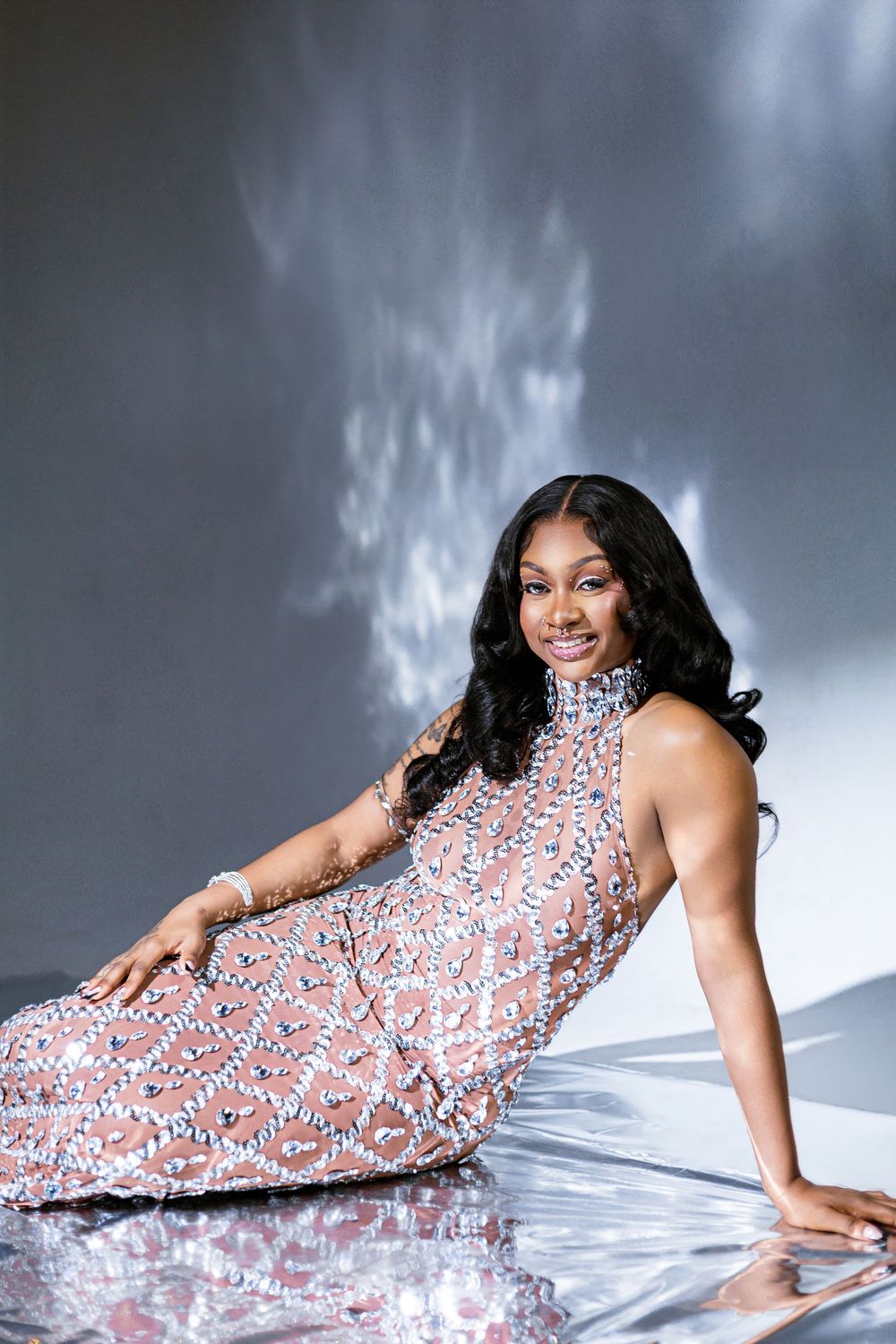A smiling woman in a sparkling sequined gown poses gracefully on a reflective floor against a gray backdrop.