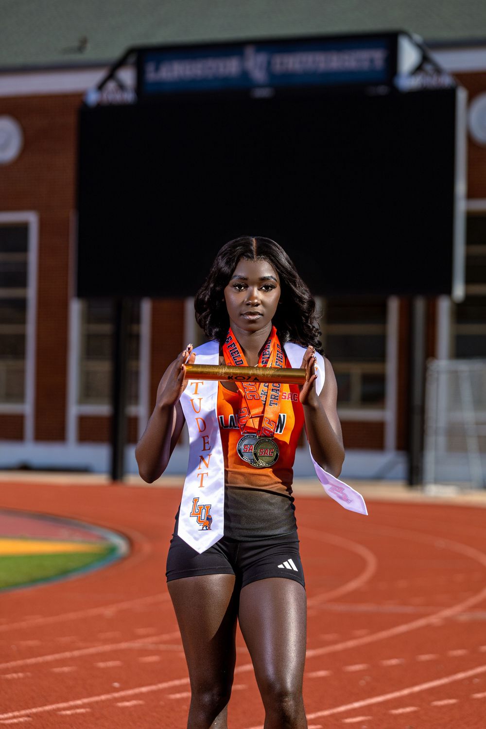 Female track athlete in orange uniform holds trophy and medal on red track at Langston University stadium.